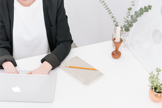 A person works at a desk on a laptop. A notepad, candle and two plants are also on the desk.